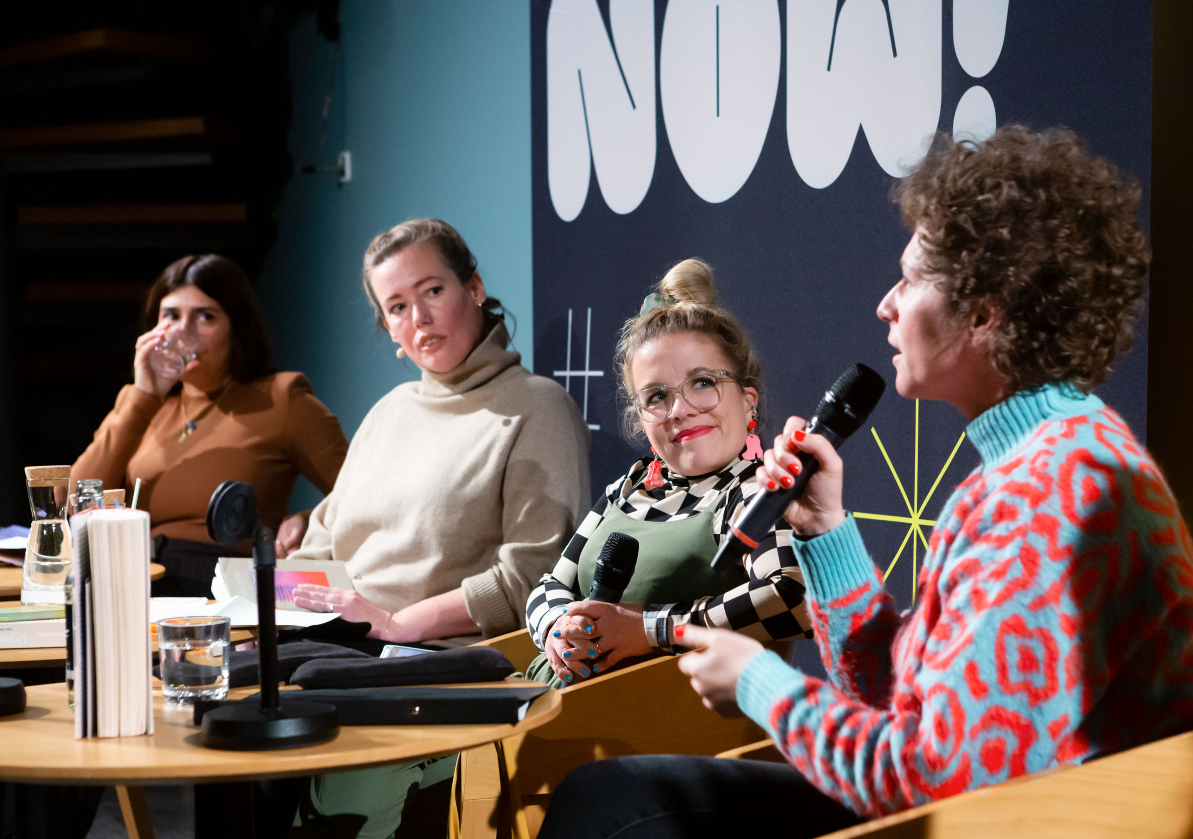 RIOT NOW! Vier Frauen, die Autorinnen, sitzen auf der Buehne vor schwarzem Hintergrund mit Lettern N O W ! Frau haelt Mikrofon in der Hand