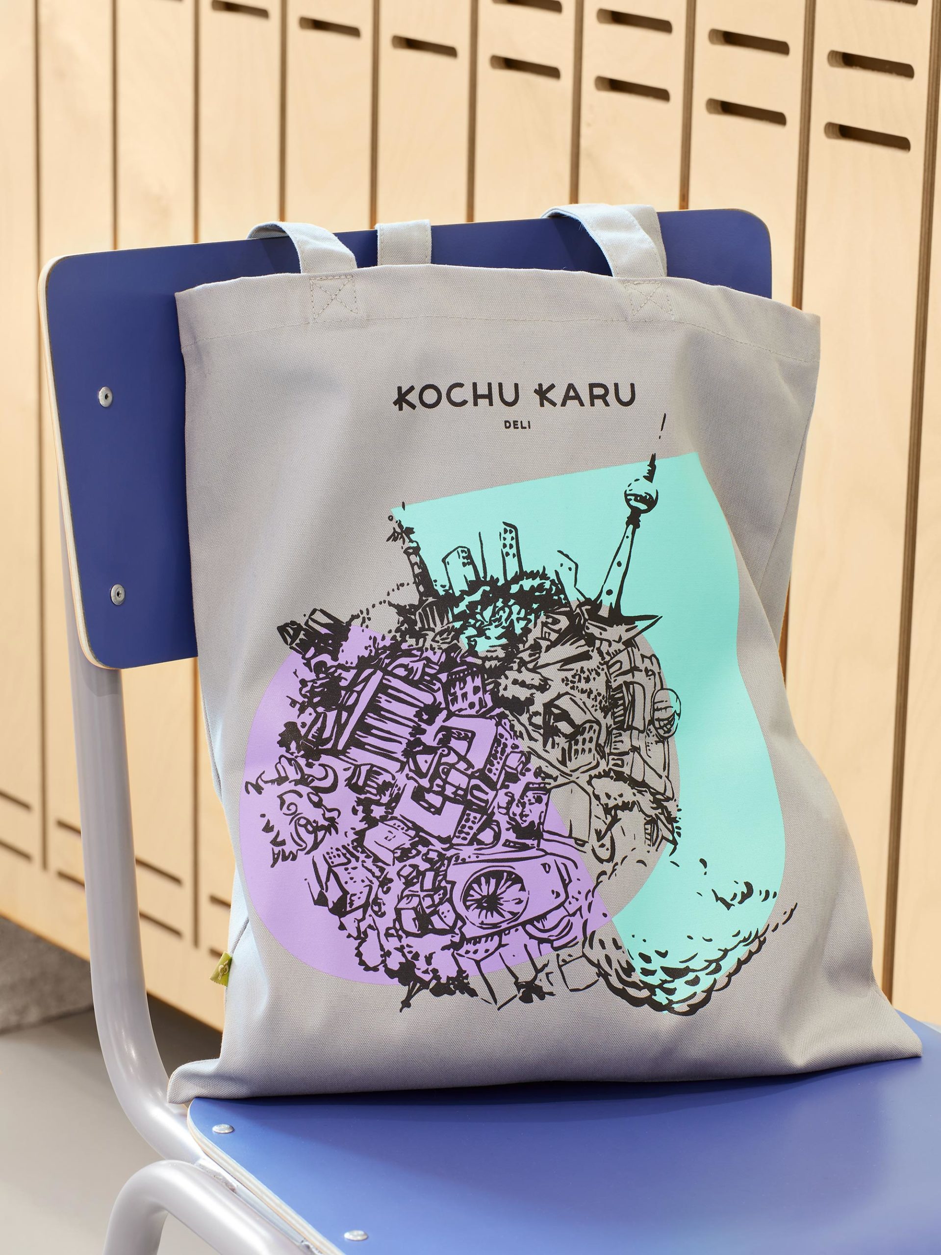 Kochu Karu Deli Stoffbeutel in grau mit farbiger Illustration und Logo auf blauem Stuhl